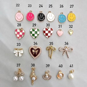 May include: A collection of twenty metal charms labeled 22 through 41, featuring various designs including smiley faces, checkered hearts, bows, pearls, and seashells in colors like pink, black, white, blue, yellow, red, and gold tones.