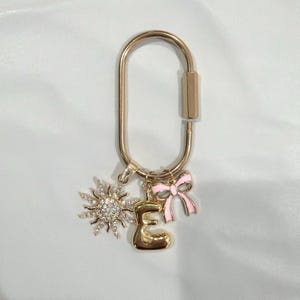 May include: Gold-tone keychain with a sunburst charm, a letter "E" charm, and a pink bow charm. The sunburst charm is encrusted with small, clear crystals. The bow charm has pink enamel and gold-tone accents. The keychain is a decorative accessory.