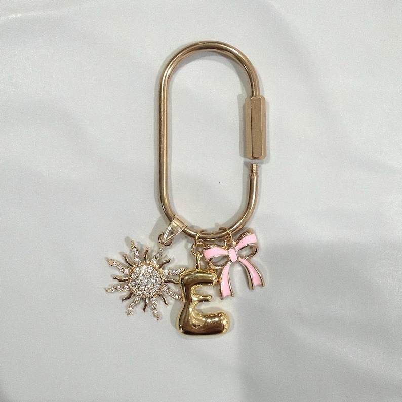 May include: Gold-tone keychain with a sunburst charm, a letter "E" charm, and a pink bow charm. The sunburst charm is encrusted with small, clear crystals. The bow charm has pink enamel and gold-tone accents. The keychain is a decorative accessory.