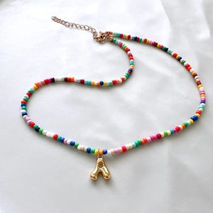 May include: A vibrant beaded necklace with a gold-coloured letter "A" pendant. The necklace features small, round beads in various colours, including red, blue, green, and yellow. The necklace has a gold-coloured chain and clasp.