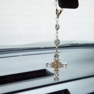 May include: A gold-coloured car charm with a cross pendant adorned with clear, sparkling crystals. The cross has a detailed central design and is suspended from a chain with crystal accents. The charm hangs from a clip.