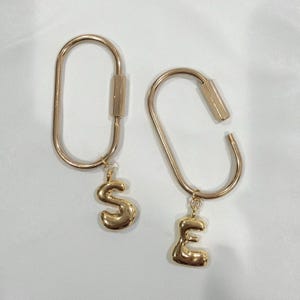 May include: Two gold-colored keychains. One keychain is closed and has a gold letter "S" charm. The other keychain is open and has a gold letter "E" charm. Both keychains are on a white background.