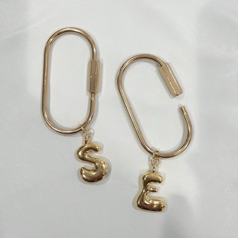 May include: Two gold-colored keychains. One keychain is closed and has a gold letter "S" charm. The other keychain is open and has a gold letter "E" charm. Both keychains are on a white background.
