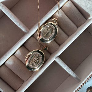K&ouml;nnte beinhalten: Zwei goldfarbene ovale Anh&auml;nger-Halsketten. Ein Anh&auml;nger zeigt ein Kreuz, der andere eine religi&ouml;se Figur. Beide Anh&auml;nger sind mit kleinen, funkelnden Steinen verziert und h&auml;ngen an zarten Ketten. Die Halsketten werden in einer Schmuckschatulle pr&auml;sentiert.