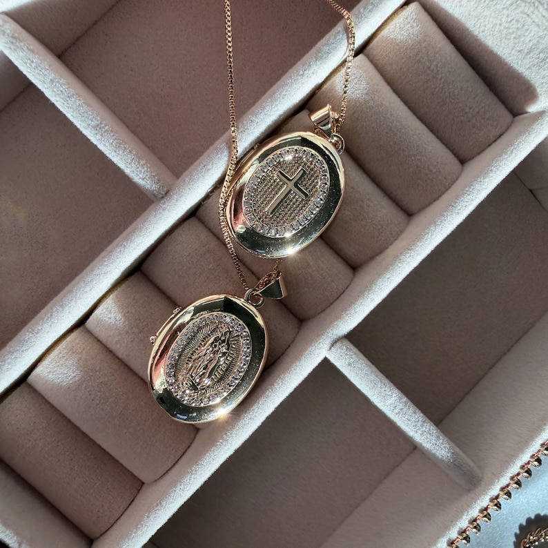K&ouml;nnte beinhalten: Zwei goldfarbene ovale Anh&auml;nger-Halsketten. Ein Anh&auml;nger zeigt ein Kreuz, der andere eine religi&ouml;se Figur. Beide Anh&auml;nger sind mit kleinen, funkelnden Steinen verziert und h&auml;ngen an zarten Ketten. Die Halsketten werden in einer Schmuckschatulle pr&auml;sentiert.