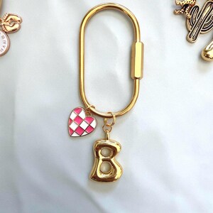 May include: A gold-toned oval carabiner keychain featuring two dangling charms. One charm is a small heart with a pink and white checkered pattern, and the other charm is a three-dimensional gold-toned letter B with a smooth, polished finish.