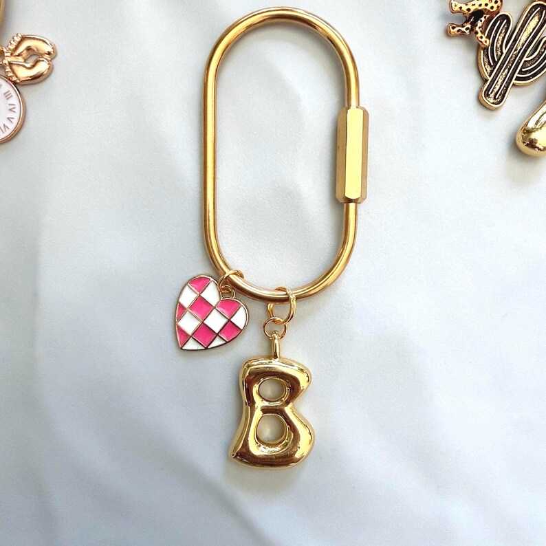May include: A gold-toned oval carabiner keychain featuring two dangling charms. One charm is a small heart with a pink and white checkered pattern, and the other charm is a three-dimensional gold-toned letter B with a smooth, polished finish.