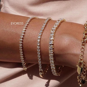 Op de afbeelding: Goudkleurige armbanden met heldere, sprankelende stenen. De armbanden hebben verschillende steensneden, waaronder rond en vierkant. Het woord "EVORESS" staat links op de afbeelding. De armbanden worden op een arm getoond.