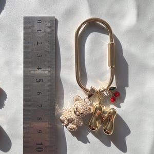 May include: Gold-toned keychain with a large oval clasp. The keychain features a small, jeweled teddy bear charm, a gold letter "M" charm, and a charm with red cherries. The keychain is shown next to a ruler.