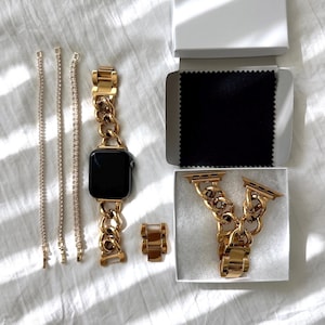 May include: A gold-colored Apple Watch with a chain-link band, a matching ring, and two rhinestone bracelets. The watch is displayed with an extra band and a black cleaning cloth in a white box. The items are arranged on a white surface.