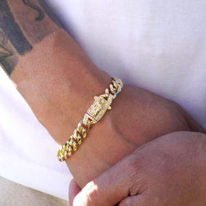 May include: A gold-colored bracelet with a cuban link chain design. The clasp is rectangular and encrusted with small, clear stones. The bracelet is worn on a wrist.
