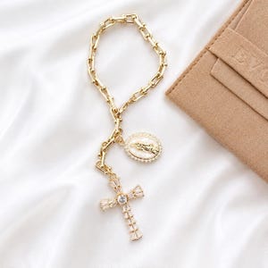 May include: A gold-toned chain bracelet featuring a rectangular link design. The bracelet includes a dangling oval pendant with a central figure surrounded by small clear stones, and a cross pendant composed of baguette-cut clear stones.