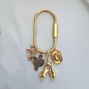 May include: A gold-toned oval carabiner keychain featuring four dangling charms: a textured leopard figure, a saguaro cactus silhouette, a smooth letter A, and a detailed cowboy hat. The metal hardware has a polished finish throughout.