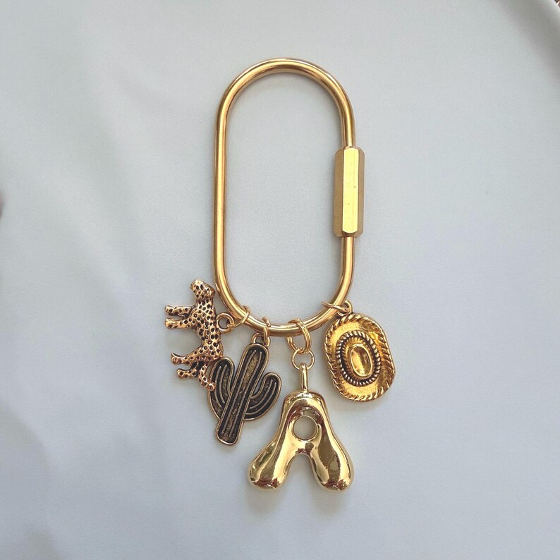 May include: A gold-toned oval carabiner keychain featuring four dangling charms: a textured leopard figure, a saguaro cactus silhouette, a smooth letter A, and a detailed cowboy hat. The metal hardware has a polished finish throughout.