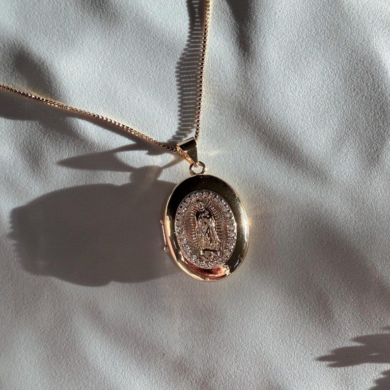 K&ouml;nnte beinhalten: Goldfarbene ovale Medaillon-Anh&auml;nger-Halskette. Das Medaillon zeigt eine religi&ouml;se Figur, umgeben von kleinen, klaren Steinen. Der Anh&auml;nger h&auml;ngt an einer zarten Kette. Die Halskette wird auf einer wei&szlig;en Oberfl&auml;che pr&auml;sentiert.