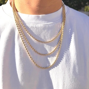 May include: Three gold-colored rope chain necklaces of varying lengths. The necklaces are worn against a white t-shirt. The chains have a twisted rope design and are displayed in a layered style.