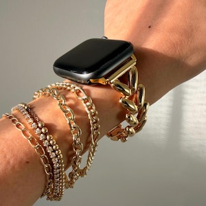 May include: A gold-toned Apple Watch with a black screen is shown on a wrist, paired with multiple gold bracelets. The bracelets include a chain link style, a beaded bracelet, and a delicate chain with small stones. The jewelry is displayed against a neutral background.