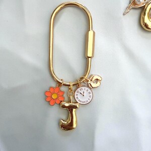 May include: A gold-toned oval carabiner keychain featuring four dangling charms: an orange flower with a yellow center, a large gold letter J, a round white clock face with black Roman numerals and hands, and a small gold-toned footprint charm.