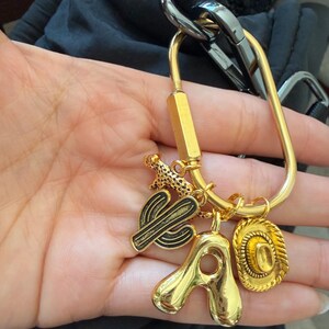 May include: A gold-toned metal carabiner keychain featuring four dangling charms: a small leopard figure, a black and gold cactus, a large polished gold letter A, and a textured gold cowboy hat. The carabiner has a screw-lock mechanism.
