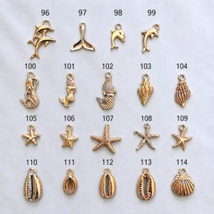 May include: A collection of gold-toned metal charms featuring nautical and marine themes, including dolphins, mermaids, starfish, and various seashells. Each charm is labeled with a number from 96 to 114, arranged in a grid on a plain background.