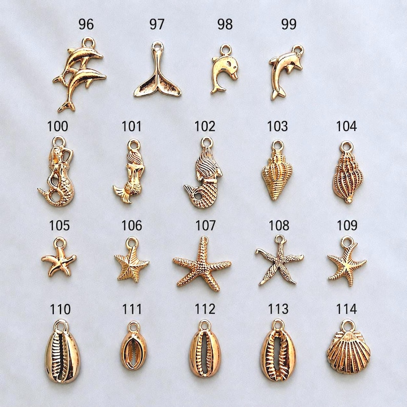 May include: A collection of gold-toned metal charms featuring nautical and marine themes, including dolphins, mermaids, starfish, and various seashells. Each charm is labeled with a number from 96 to 114, arranged in a grid on a plain background.