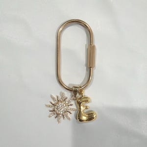 May include: Gold-colored keychain with a sun charm and an initial "E" charm. The sun charm is encrusted with small, clear crystals. The keychain has a simple, oval shape with a clasp closure.