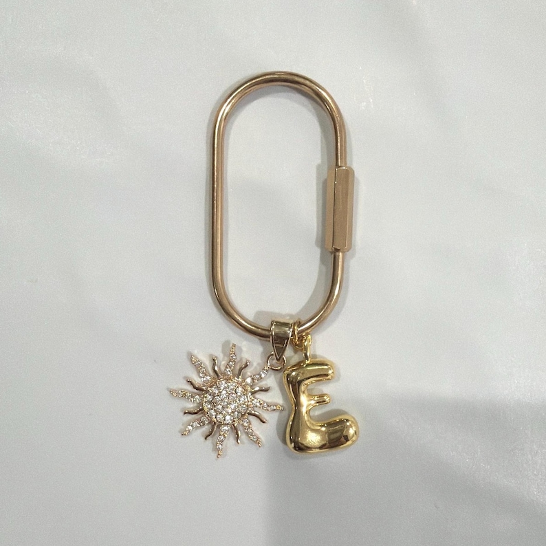 May include: Gold-colored keychain with a sun charm and an initial "E" charm. The sun charm is encrusted with small, clear crystals. The keychain has a simple, oval shape with a clasp closure.