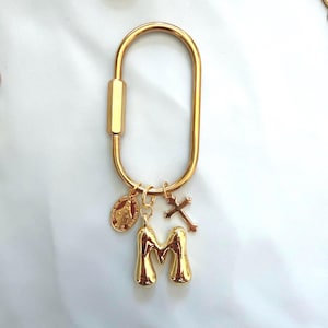 May include: A gold-toned oval carabiner keychain featuring a polished finish. Attached to the ring are three charms: a puffy, rounded letter M, a small oval religious medal depicting a figure, and a slender, detailed cross pendant in matching gold.