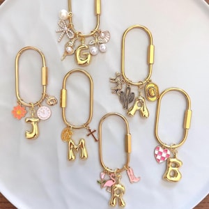 May include: Six gold-toned oval carabiner keychains, each featuring a unique set of charms. Charms include letters G, J, M, R, B, and A, alongside various motifs like pearls, flowers, a clock, a cross, a cactus, a cowboy hat, a flamingo, and a heart.