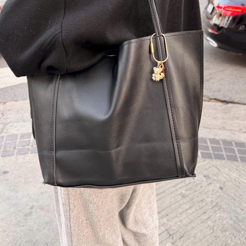 May include: A black tote bag with a gold-colored charm shaped like a bear. The bag is made of a smooth material and has a simple, rectangular design. The charm has red and white accents.