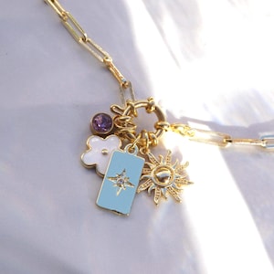 May include: Gold-toned necklace with a paperclip chain and a variety of charms. Charms include a purple gemstone, a white flower, a light blue rectangular charm with a starburst design, and a sunburst charm.