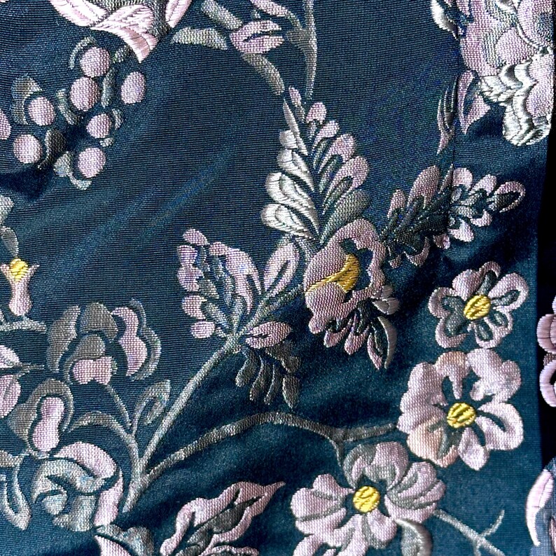 Pu&ograve; includere: Primo piano di un tessuto blu scuro con un intricato motivo floreale. Il design presenta fiori, foglie e steli lilla chiaro e grigio argento, con accenti gialli. Il tessuto ha una leggera lucentezza.