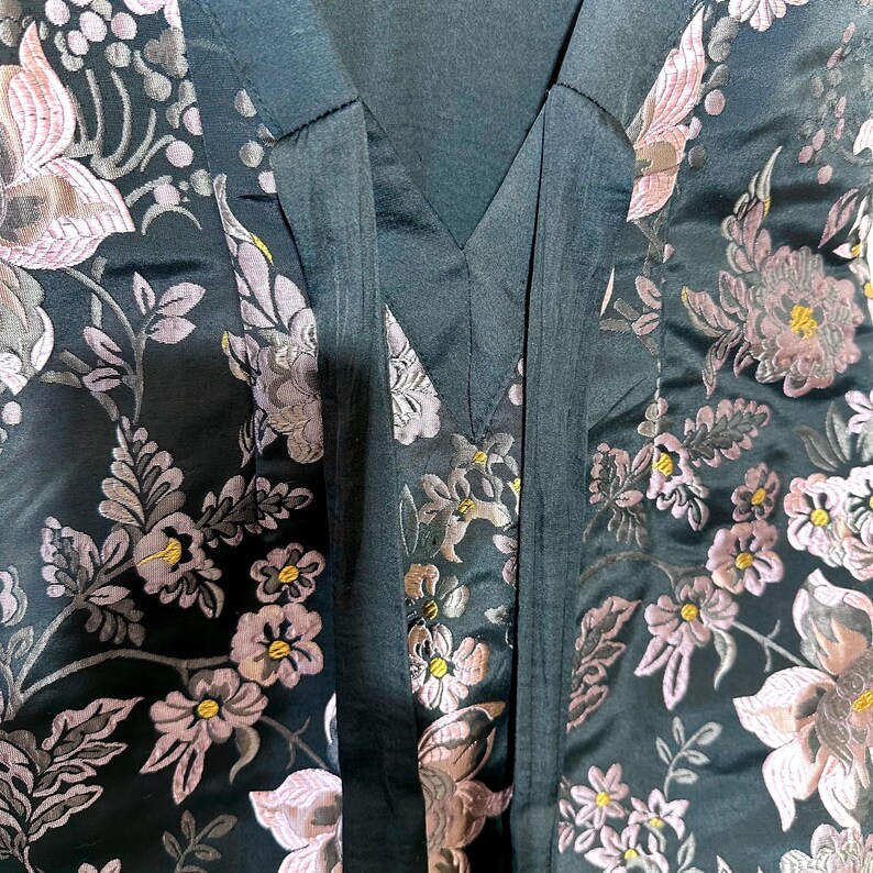 Pu&ograve; includere: Primo piano di un tessuto verde scuro con un motivo floreale. Il design presenta fiori rosa e grigi con centri gialli e foglie verdi. Il tessuto ha una lucentezza satinata ed &egrave; probabilmente utilizzato per abbigliamento o arredamento.