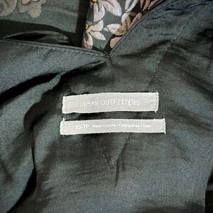 Pu&ograve; includere: Primo piano di un capo di abbigliamento verde scuro con un motivo floreale. Il capo ha un'etichetta che recita "URBAN OUTFITTERS" e "XS-TP Made in China / Fabriqu&eacute; en Chine". Il tessuto sembra essere raso o un materiale simile.