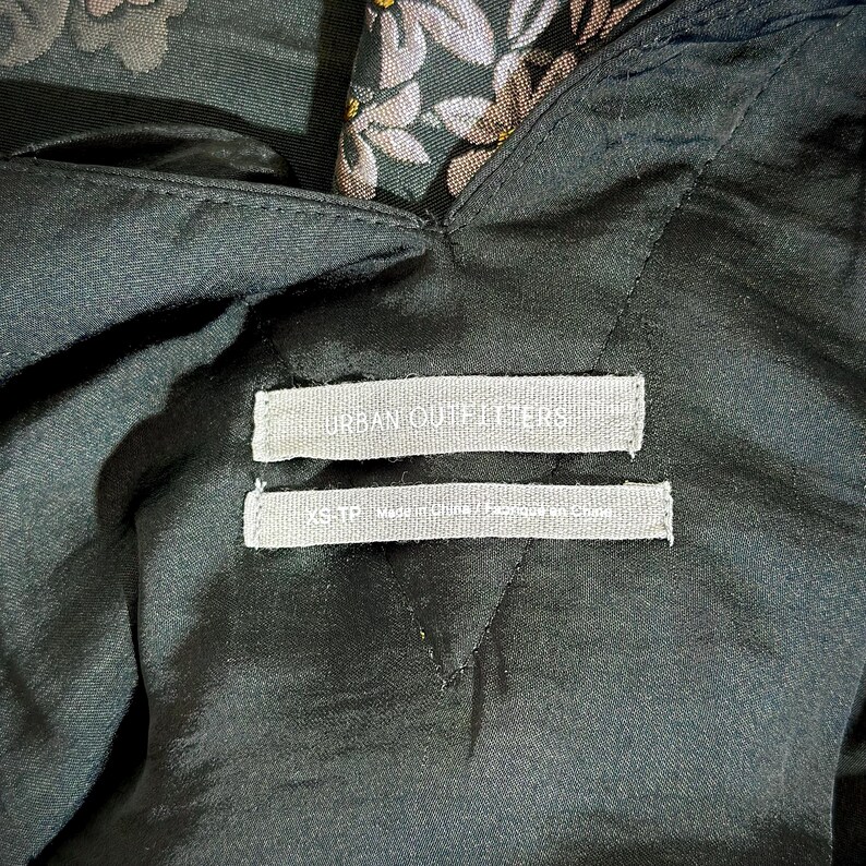 Pu&ograve; includere: Primo piano di un capo di abbigliamento verde scuro con un motivo floreale. Il capo ha un'etichetta che recita "URBAN OUTFITTERS" e "XS-TP Made in China / Fabriqu&eacute; en Chine". Il tessuto sembra essere raso o un materiale simile.