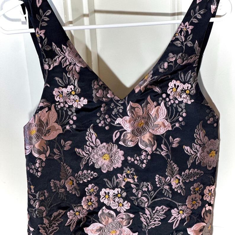 Pu&ograve; includere: Top senza maniche nero con scollo a V, ornato da un motivo floreale rosa e oro. I fiori sono dettagliati, con foglie e steli. Il tessuto ha una leggera lucentezza. Il top &egrave; appeso a una gruccia bianca.