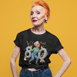 Op de afbeelding: Een zwart t-shirt met korte mouwen voorzien van een grafische illustratie van een ouder persoon met wit haar, een zonnebril, een groen shirt met lange mouwen en een blauwe spijkerbroek, die een rock-and-roll handgebaar maakt voor het getal 80.