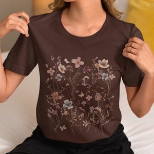 May include: A brown t-shirt featuring a floral design with various flowers in shades of pink, white, and blue. The shirt has a crew neck and short sleeves. The word "Brown" is at the bottom of the image.