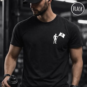 以下が含まれることがあります： 左胸に小さな白いグラフィックが施された黒の半袖Tシャツ。グラフィックには、ドクロと骨が交差したデザインの旗を持つ人物が立っている様子が描かれています。右上の円形のバッジにはBLACKと書かれています。