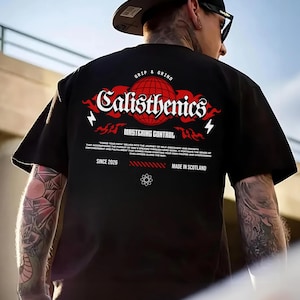 Può includere: Una t-shirt nera con una grafica a globo rosso e testo bianco che recita Calisthenics, Grip & Grind, Mastering Control e Made in Scotland. Il design include piccoli fulmini, accenti di fiamma e un paragrafo di testo.