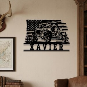 Könnte beinhalten: Schwarze Metall-Wandkunst mit einem Offroad-Fahrzeug-Design, einem amerikanischen Flaggenhintergrund und dem Namen "XAVIER" darunter. Das Kunstwerk enthält detaillierte Silhouetten von Bäumen und Fahrzeugmerkmalen.