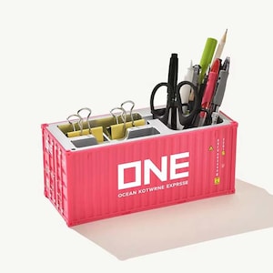 Può includere: Un organizer da scrivania rosa a forma di container con il testo ONE OCEAN KOTWRNE EXPRSSE stampato sul lato. Il contenitore presenta diversi scomparti che contengono clip gialle, forbici nere e varie penne e matite per l'ufficio.