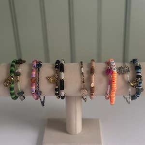 Puede incluir: Exhibición de pulseras de cuentas coloridas. Las pulseras presentan varios diseños, incluyendo dijes dorados, cuentas de letras y una mezcla de colores como verde, rosa, negro y blanco. Están dispuestas sobre un expositor de color crema.