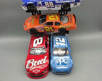Könnte beinhalten: Vier Modell-NASCAR-Rennwagen in einer Präsentation. Die Autos sind blau, orange, rot und blau, jeweils mit Sponsorenlogos und Rennnummern. Die Nummern sind "88", "21" und "12".