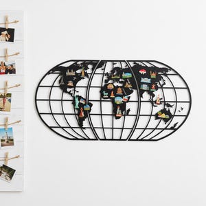Può includere: Una decorazione da parete a mappa del mondo in metallo nero con un design a griglia. La mappa presenta calamite in miniatura colorate di monumenti, tra cui la Torre Eiffel e la Statua della Libertà. La mappa è montata su una parete bianca.