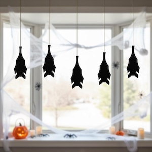 May include: Five black bat decorations hanging from thin strings in front of a window. The window is decorated with white spiderwebs and small plastic spiders. A lit jack-o'-lantern and candles sit on the windowsill, creating a Halloween theme.