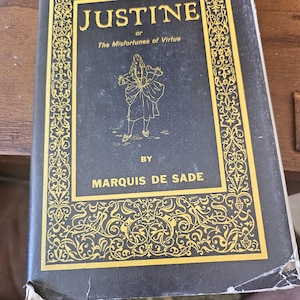 Puede incluir: Un libro antiguo titulado "Justine o Los Infortunios de la Virtud" de Marquis de Sade. La portada es negra con letras doradas y bordes decorativos. Una ilustración dorada de una persona está en el centro de la portada.