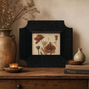 May include: A rectangular picture frame with a textured black finish containing a botanical print of red poppies. The frame features a unique notched corner design and is displayed alongside a large ceramic vase, a candle, and two books.