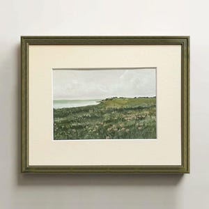 May include: A framed landscape painting featuring a grassy field with small pink flowers, a distant coastline, and a cloudy sky. The artwork is enclosed in a textured olive green frame with a wide, off-white mat board surrounding the central image.