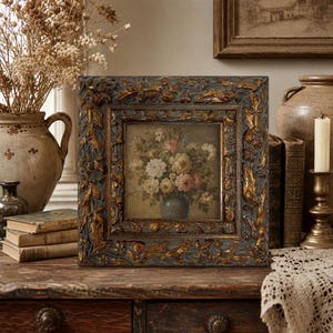 May include: Square framed floral painting with a dark blue and gold ornate frame. The painting depicts a bouquet of white and pink flowers in a blue vase. The frame has a detailed, textured design.